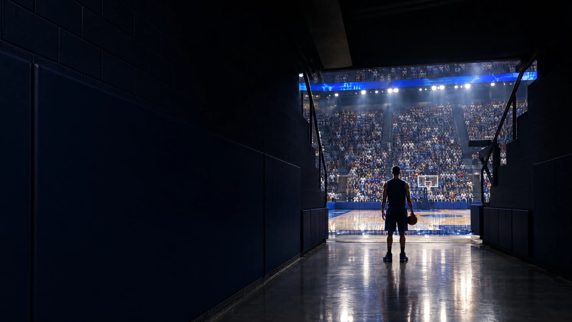 A college basketball player seen from behind at the end of a dark arena tunnel, looking out at a packed, brightly lit arena